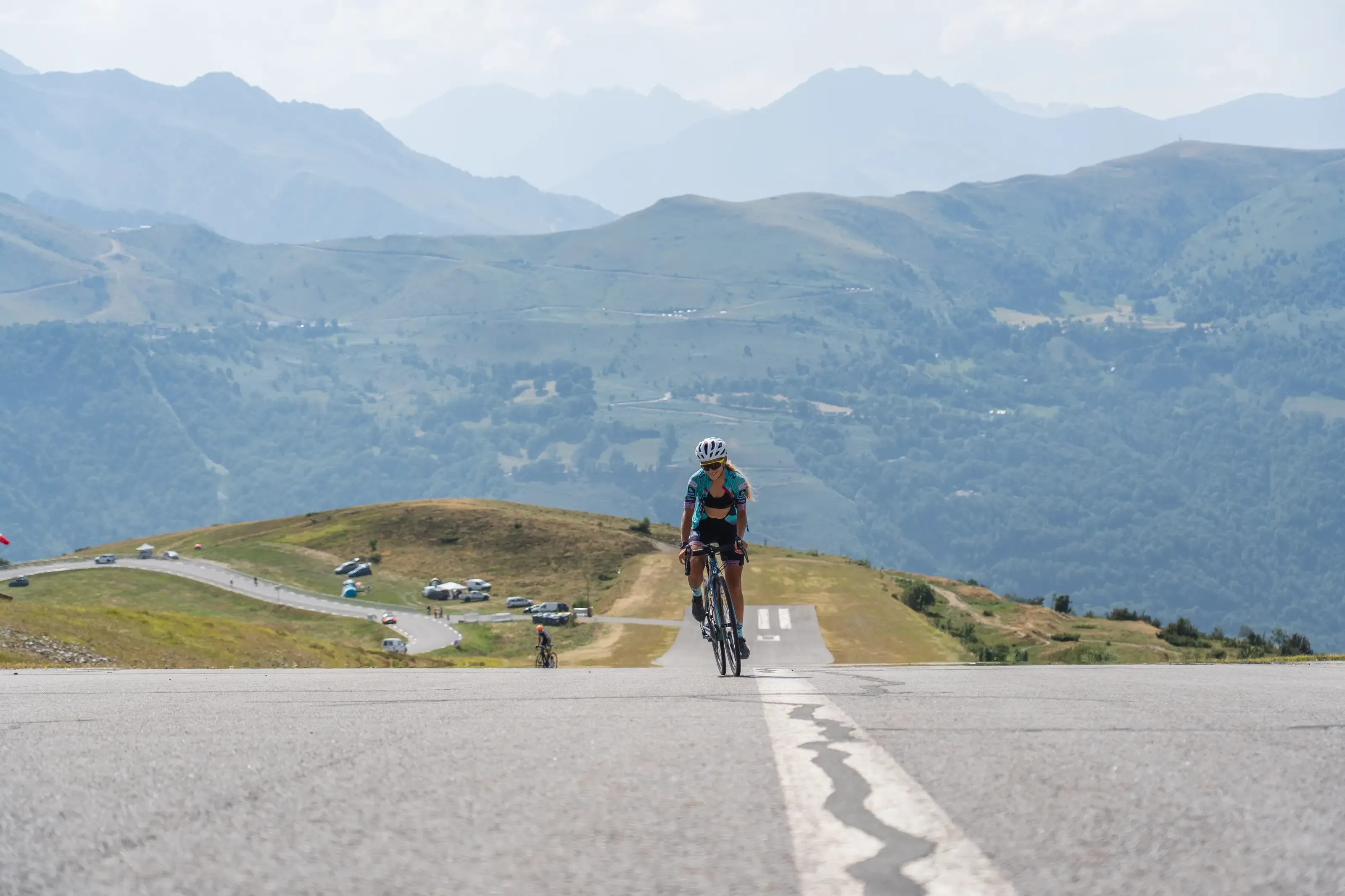 Une cycliste seule gravit à vélo une piste d'altiport en pente, face à un vaste panorama de montagnes et de sommets à l'horizon.
